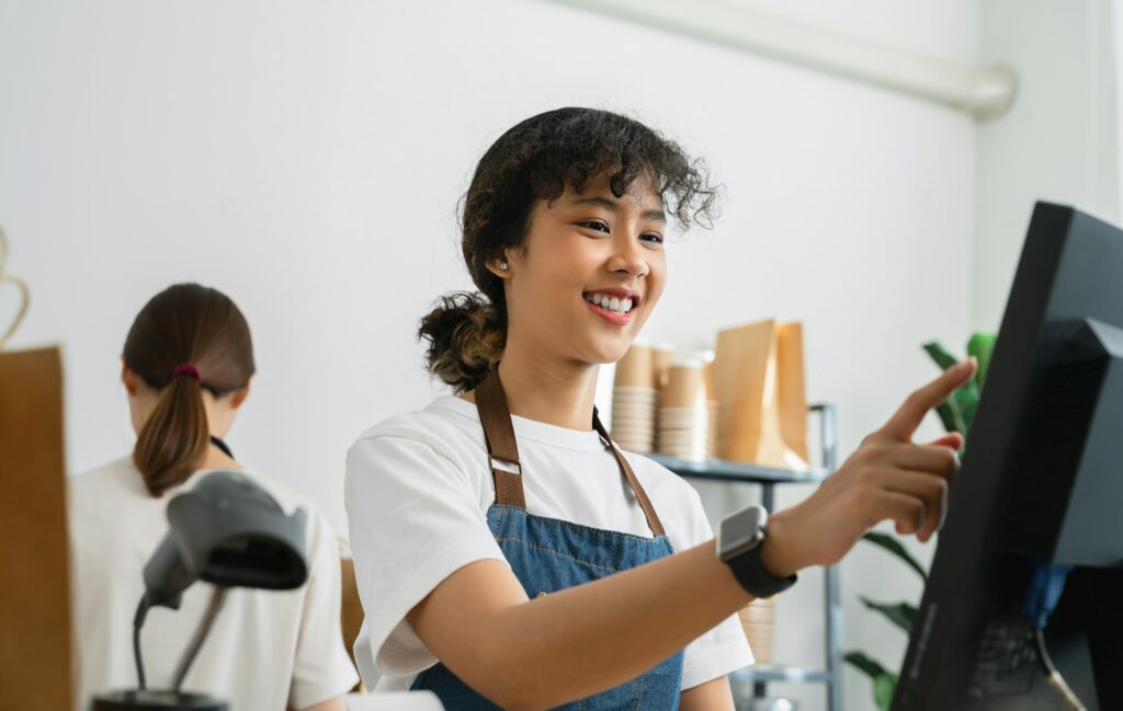 woman cashier wears an apron and using pos terminal to input orders on coffee shop counter.